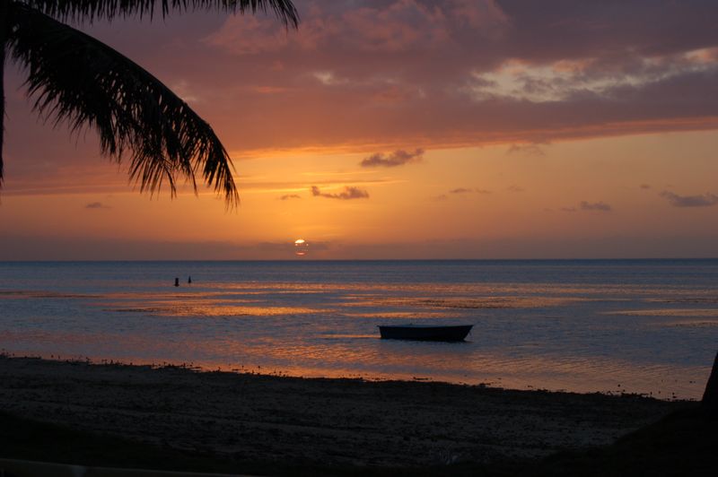 An orange sunset over the calm lagoon on Saipan, framed by a palm frond, with a small boat on the water